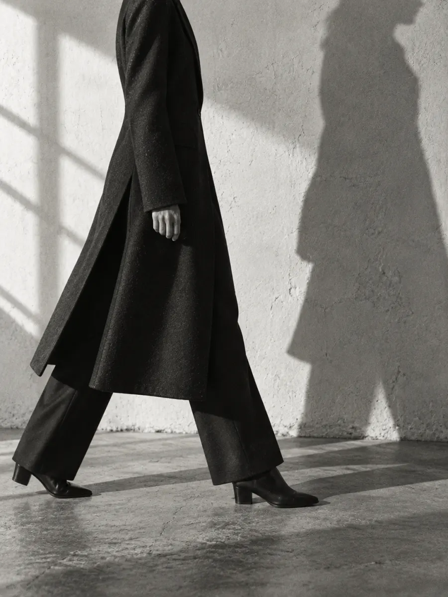 A figure in a long black coat walking past a sunlit wall